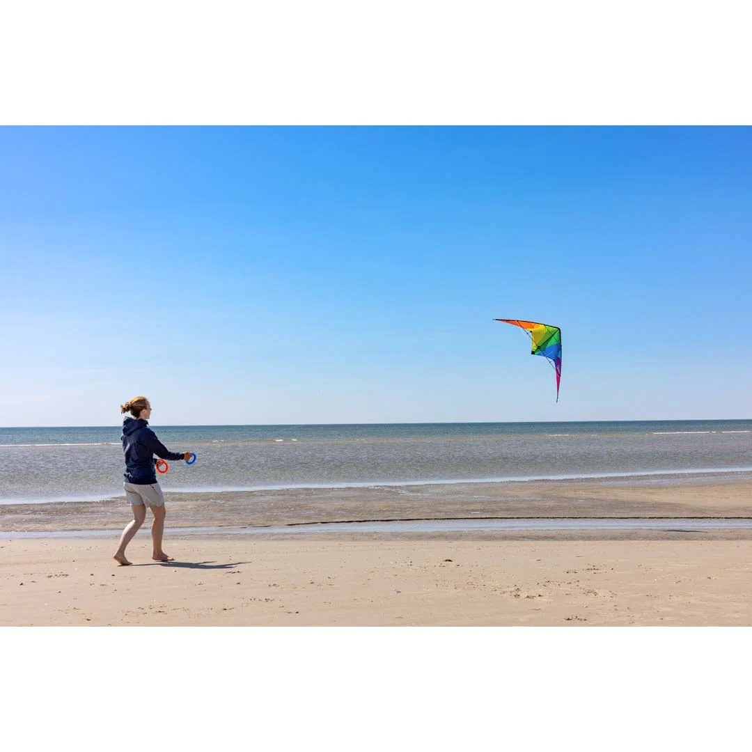 Oseba spušča akrobatskega zmaja z mavričnim vzorcem na peščeni plaži ob morju pod modrim nebom.